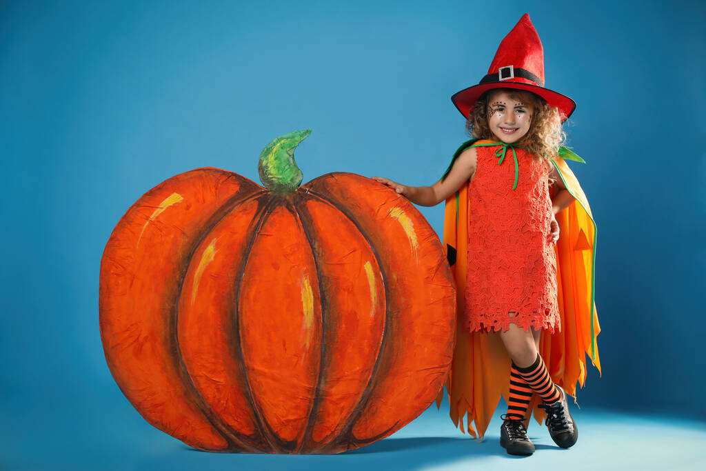 Nettes kleines Mädchen mit dekorativem Kürbis im Halloween-Kostüm auf hellblauem Hintergrund - Foto, Bild