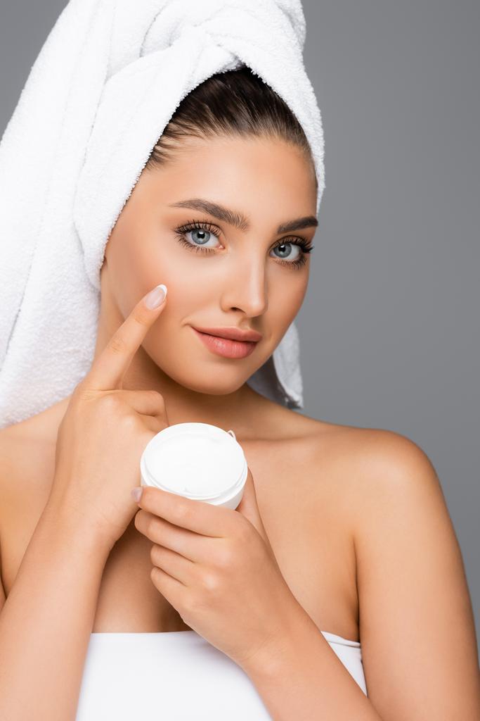 femme avec serviette sur la tête appliquer crème cosmétique sur le visage isolé sur gris - Photo, image
