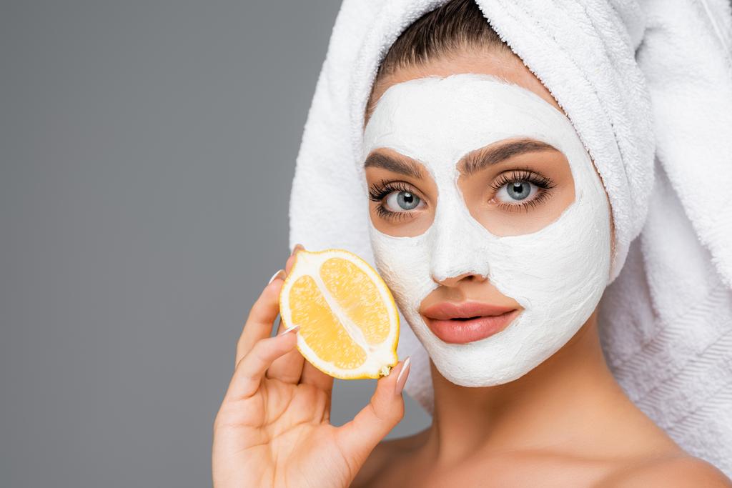 vrouw met handdoek op hoofd en klei masker op gezicht bedrijf citroen geïsoleerd op grijs - Foto, afbeelding