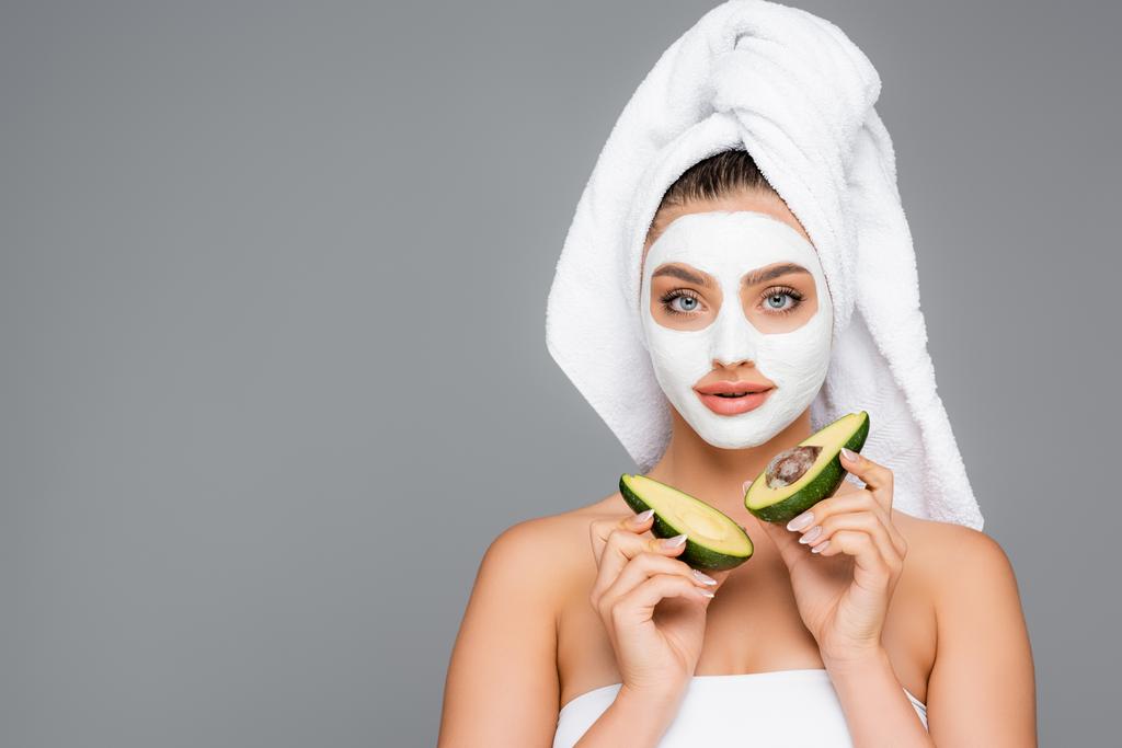 kobieta z ręcznikiem na głowie i glinianą maską na twarzy trzymając połówki awokado izolowane na szaro - Zdjęcie, obraz