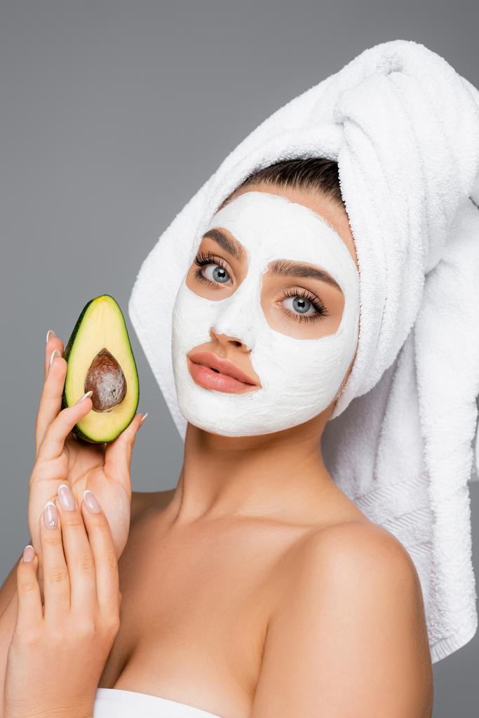 женщина с полотенцем на голове и глиняная маска на лице держа авокадо изолированы на сером - Фото, изображение