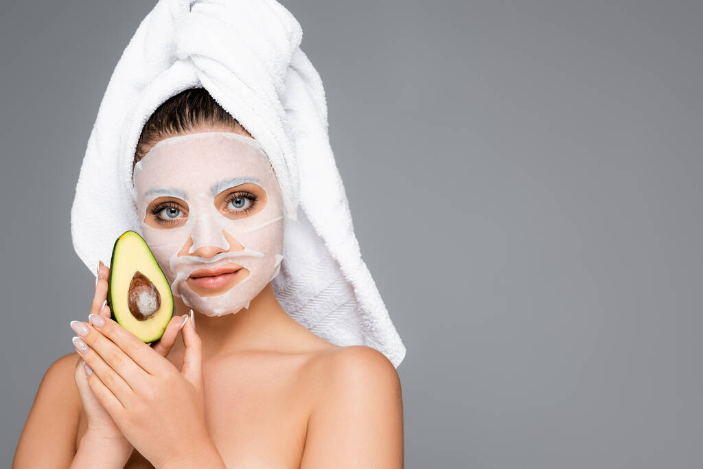 Frau mit Handtuch auf dem Kopf und Maske Laken im Gesicht hält Avocado isoliert auf grau - Foto, Bild