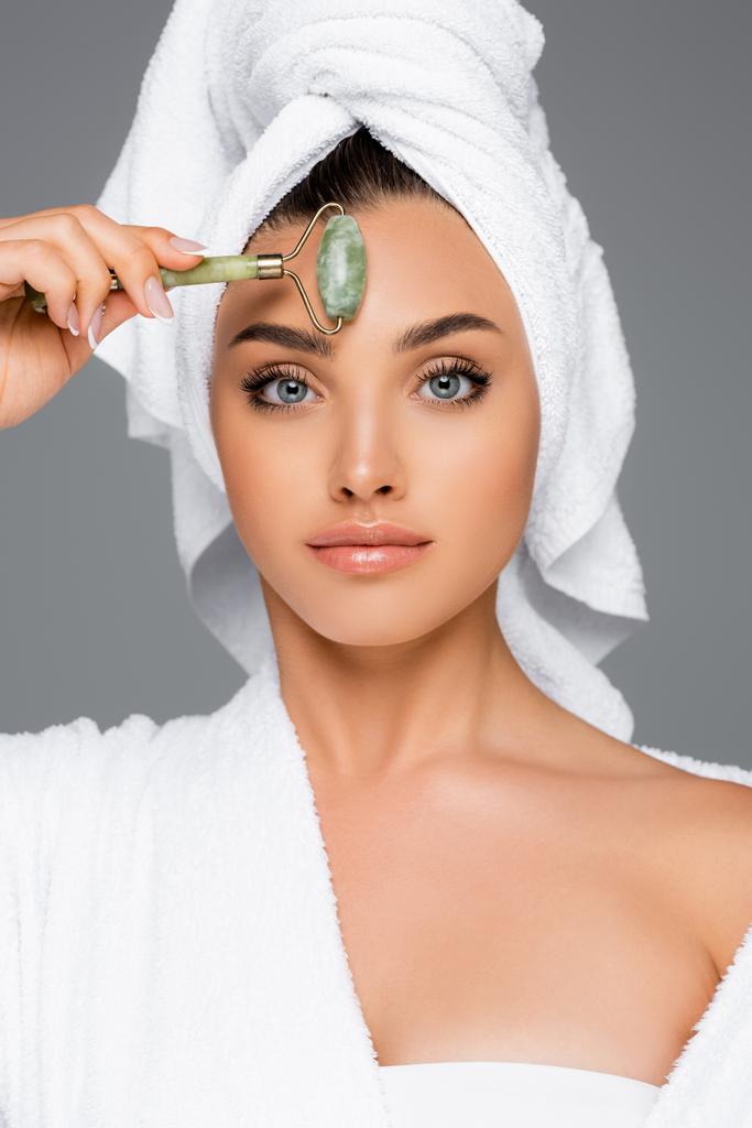 woman with towel on head using jade roller on face isolated on grey - Photo, Image