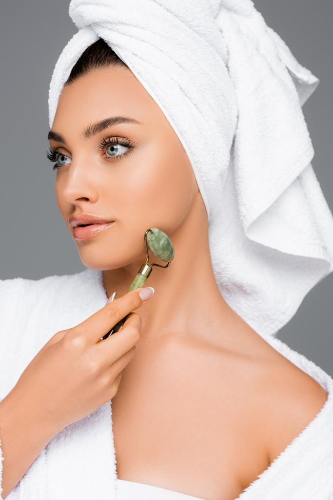 γυναίκα με πετσέτα στο κεφάλι χρησιμοποιώντας νεφρίτη κυλίνδρου στο πρόσωπο απομονώνονται σε γκρι - Φωτογραφία, εικόνα