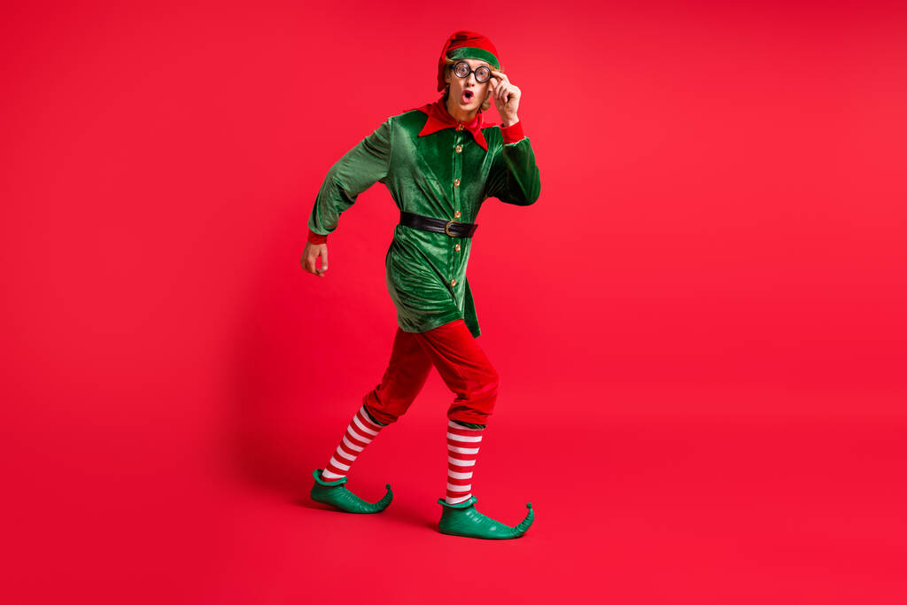 Πλήρης εικόνα προφίλ σώματος του έκπληκτου ξωτικού πάει κενό χώρο φορούν πράσινο κοστούμι απομονώνονται σε κόκκινο χρώμα λάμψη φόντο - Φωτογραφία, εικόνα