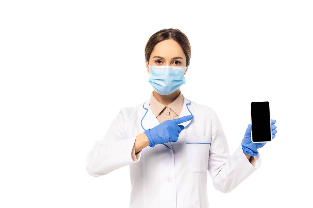Врач в медицинской маске и латексных перчатках, указывающих пальцем на смартфон, изолированный на белом - Фото, изображение
