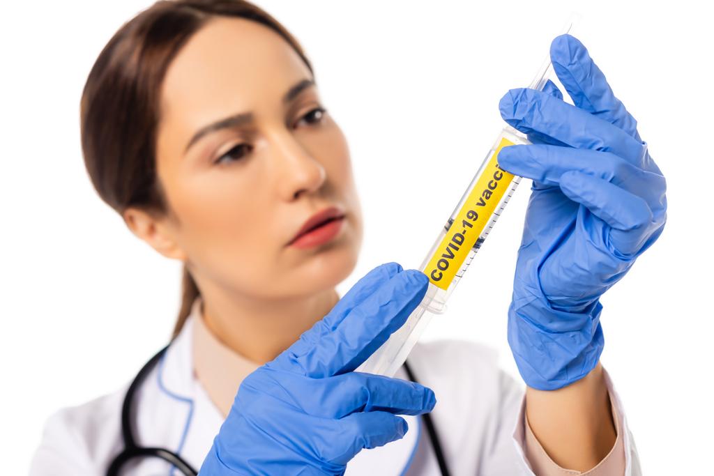Selektivní zaměření lékaře na latexové rukavice držící injekční stříkačku s kovid-19 očkovacími písmeny izolovanými na bílém - Fotografie, Obrázek