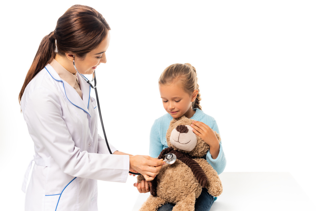 Sonriente médico examinando con estetoscopio juguete suave cerca de chica aislada en blanco - Foto, Imagen