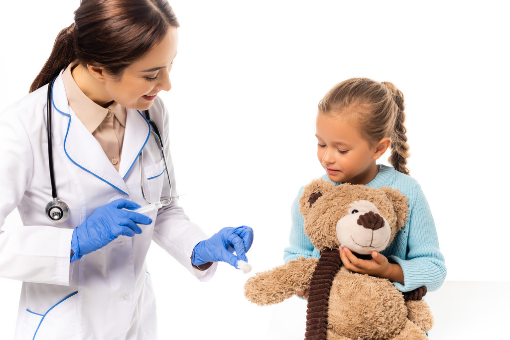 Arzt in Latex-Handschuhen hält Baumwolle und Spritze in der Nähe von Mädchen mit Teddy-Beat isoliert auf weiß - Foto, Bild