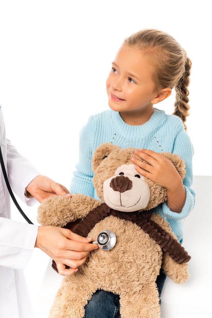 Smiling child holding soft toy near doctor with stethoscope isolated on white - Photo, Image