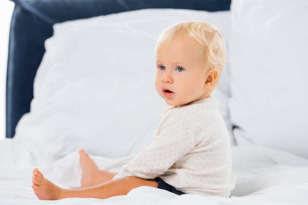 Foco seletivo do menino olhando para longe enquanto sentado na cama no fundo branco  - Foto, Imagem
