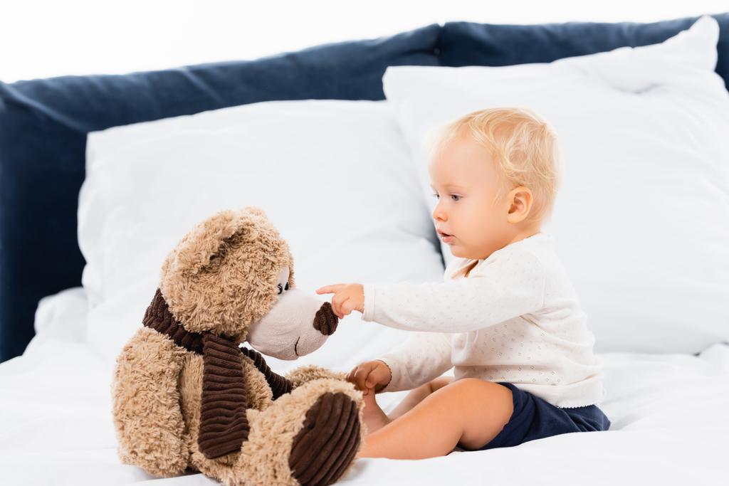Niño pequeño señalando juguete suave en la cama sobre fondo blanco  - Foto, imagen