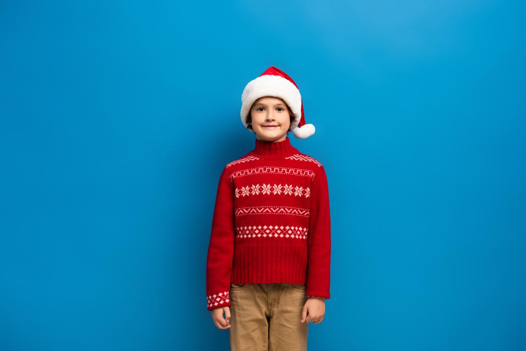happy boy in santa hat and red knitted sweater looking at camera on blue - Photo, Image