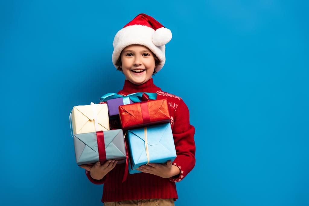 радісний хлопчик в капелюсі Санта і светрі тримає подарунки на синьому
 - Фото, зображення