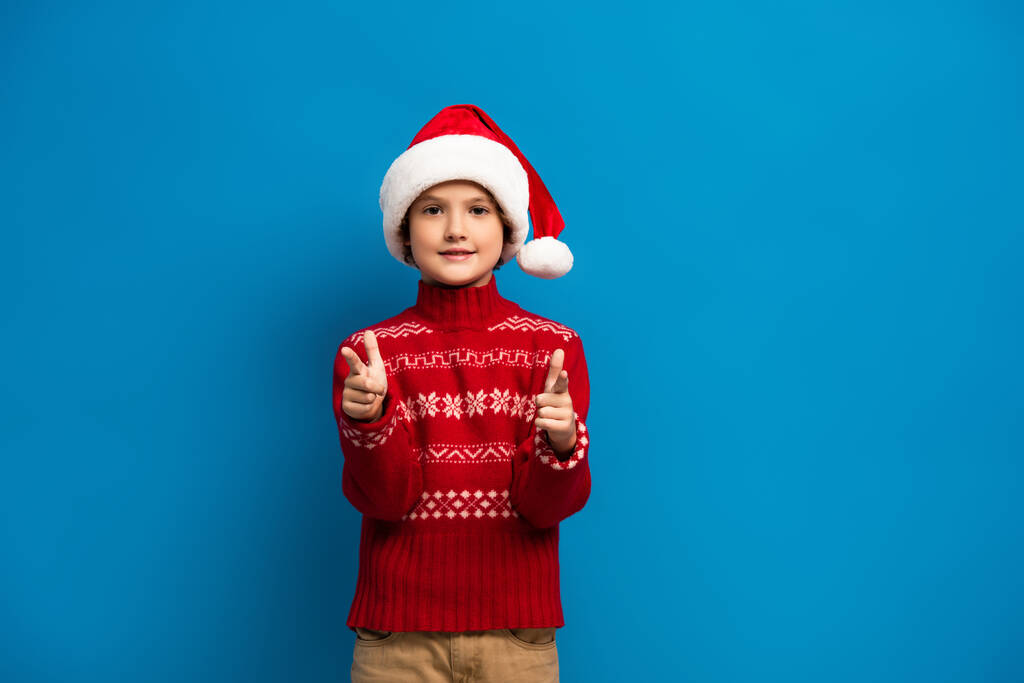 vreugdevolle jongen in kerstmuts en rode trui wijzend met vingers terwijl hij naar de camera op blauw kijkt - Foto, afbeelding