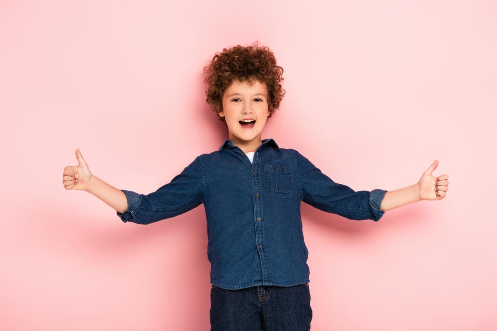 χαρούμενο και σγουρό αγόρι με απλωμένα χέρια που δείχνει τους αντίχειρες πάνω στο ροζ  - Φωτογραφία, εικόνα