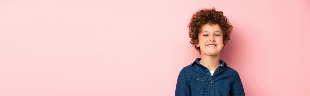 Panoramaaufnahme eines zufriedenen und lockigen Jungen im Jeanshemd, der auf rosa lächelt  - Foto, Bild
