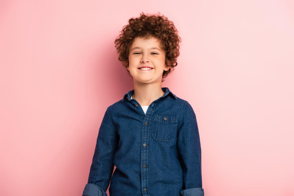 heureux et bouclé garçon en denim chemise souriant sur rose  - Photo, image