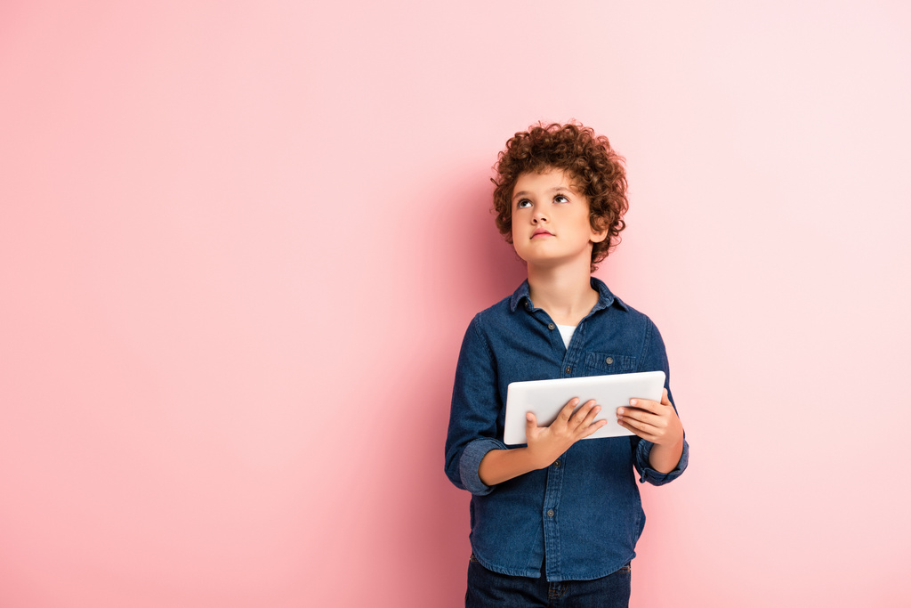kudrnatý chlapec v džínové košili vzhlíží a drží digitální tablet na růžové  - Fotografie, Obrázek