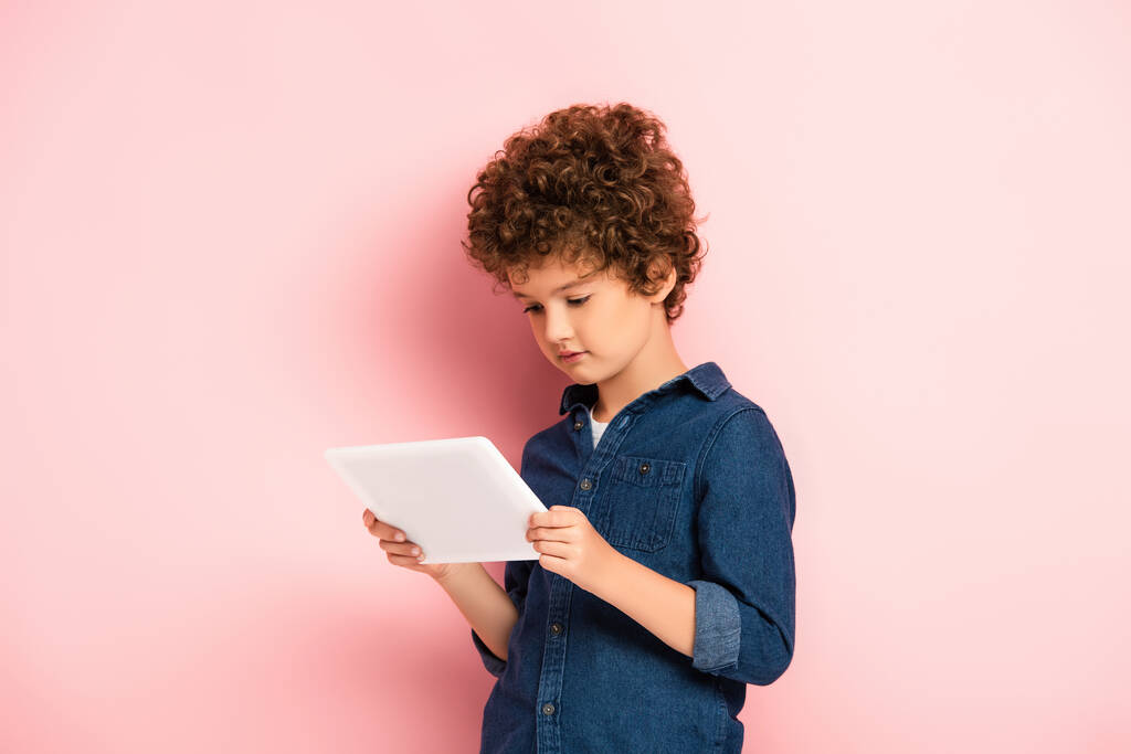 кудрявый мальчик в джинсовой рубашке смотрит на цифровой планшет на розовый  - Фото, изображение