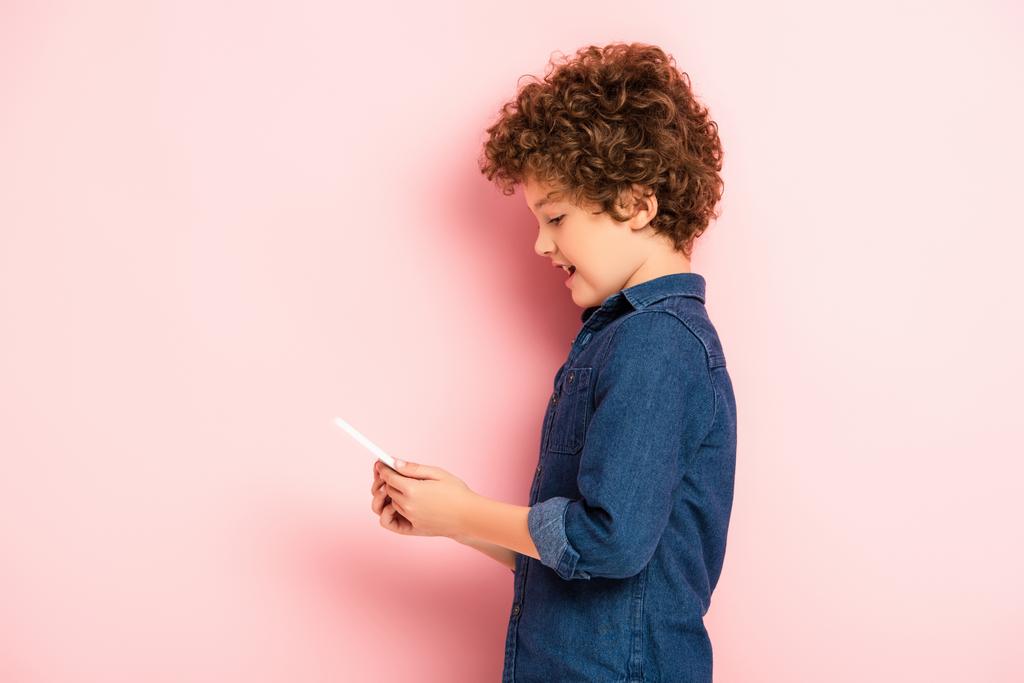 ピンクのスマートフォンを持ったデニムシャツの男の子の喜びの側面図 - 写真・画像