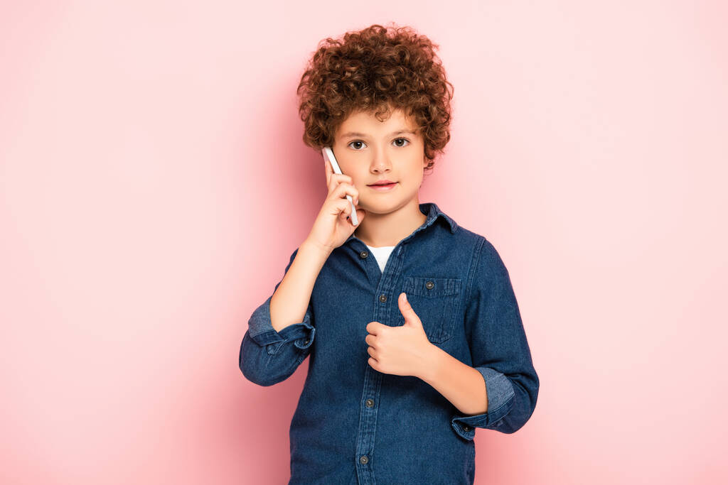 kudrnatý chlapec v džínové košili mluví na smartphone a ukazující palec nahoru na růžové - Fotografie, Obrázek