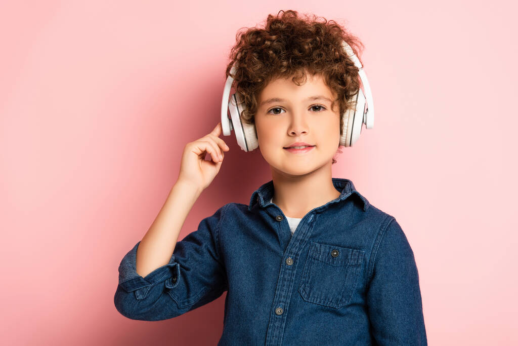 χαρούμενο και σγουρό αγόρι ακούει μουσική και αγγίζει ασύρματα ακουστικά σε ροζ  - Φωτογραφία, εικόνα