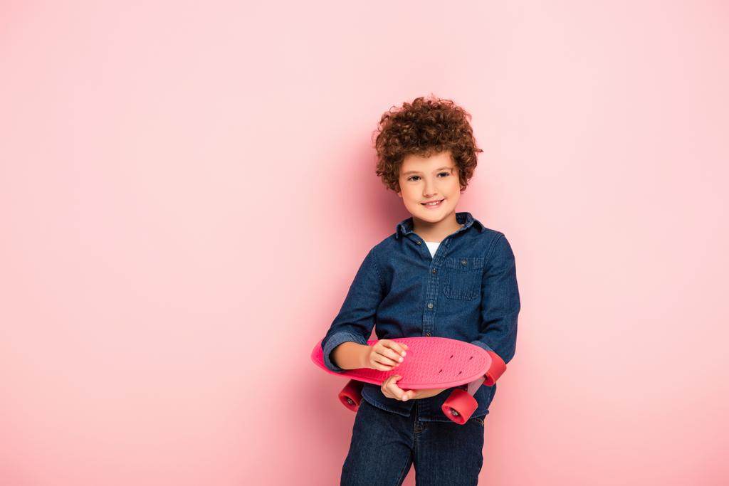 tevreden jongen holding penny board en glimlachen op roze  - Foto, afbeelding