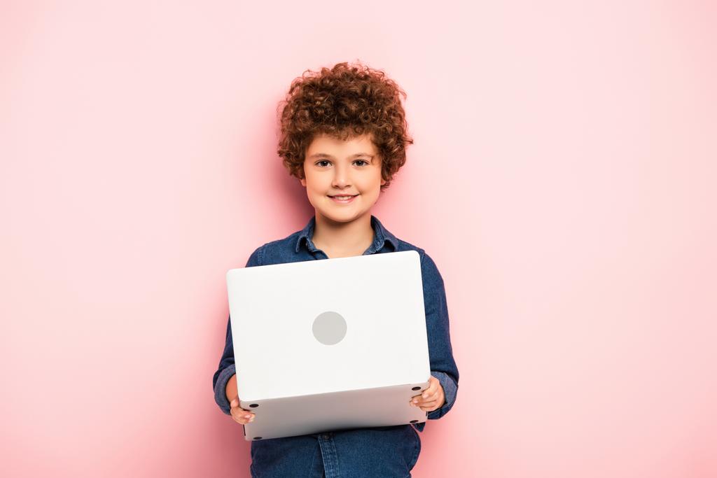 веселый и кудрявый мальчик держит ноутбук на розовом  - Фото, изображение