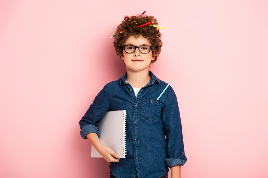 elégedett gyerek szemüvegben színes ceruzák göndör haj gazdaság jegyzettömb rózsaszín  - Fotó, kép