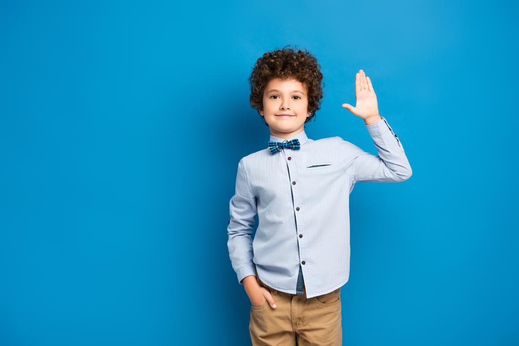 radosne dziecko w koszuli i muszce stojące z ręką w kieszeni i machającą ręką na niebieskim - Zdjęcie, obraz