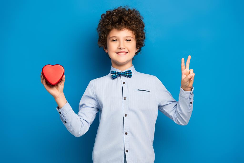 Радостный мальчик держит красную коробку формы сердца и показывает знак мира на голубом - Фото, изображение