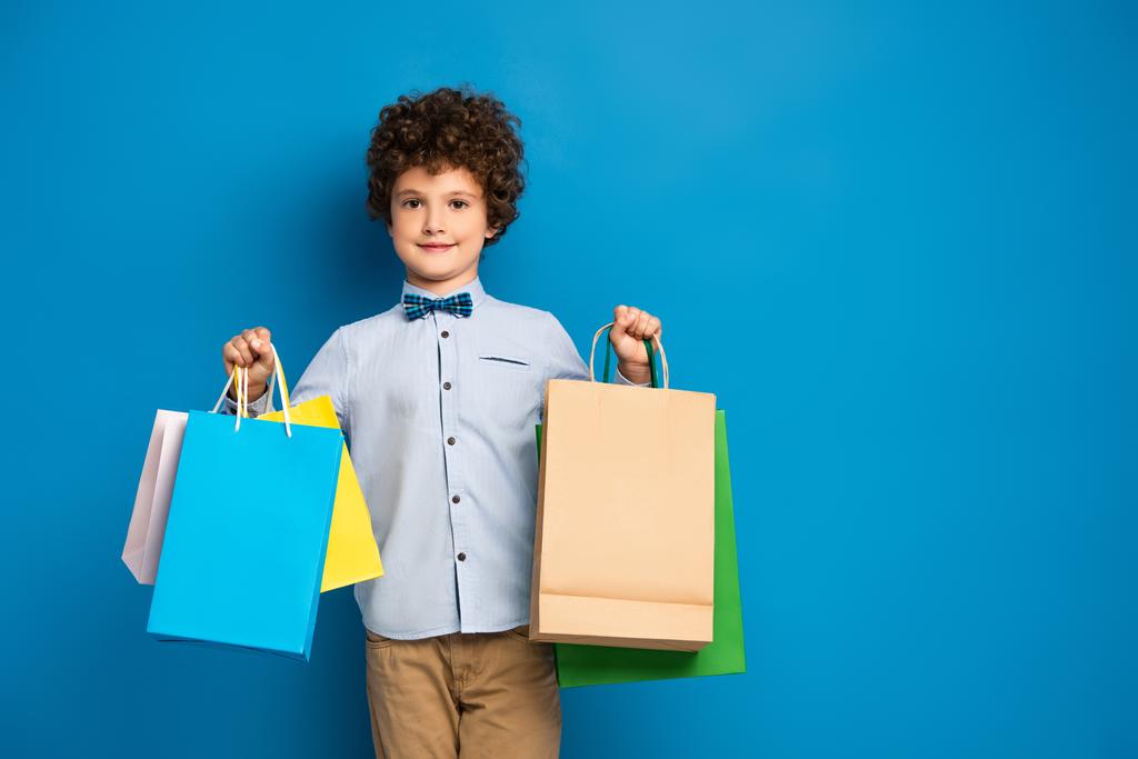 vrolijk en krullend jongen houden boodschappentassen op blauw  - Foto, afbeelding