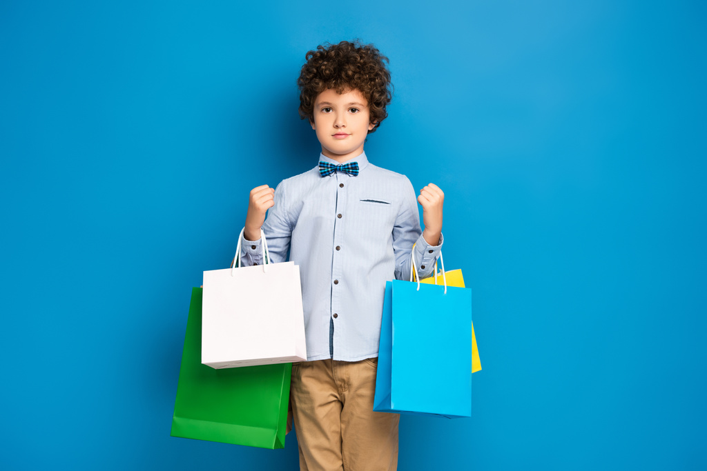 chico rizado sosteniendo bolsas de compras y de pie en azul  - Foto, Imagen