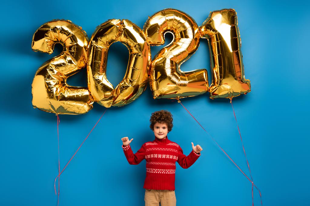 göndör fiú piros pulóverben mutatja hüvelykujját közel arany lufik 2021 számok kék - Fotó, kép
