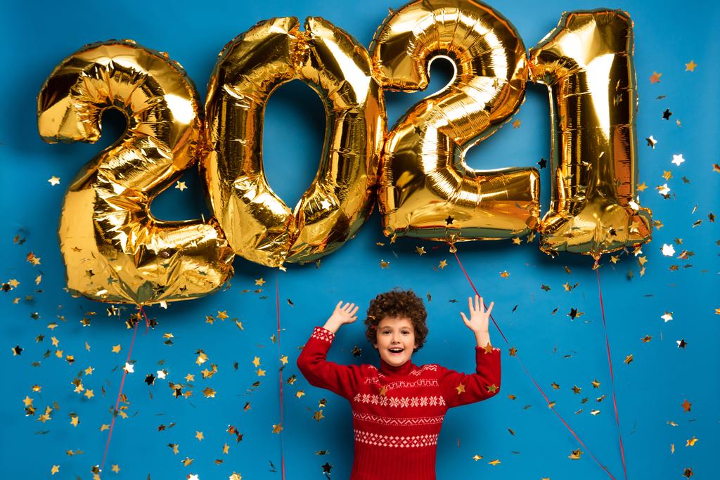 excited boy in red sweater gesturing near golden balloons with 2021 numbers and confetti on blue - Photo, Image