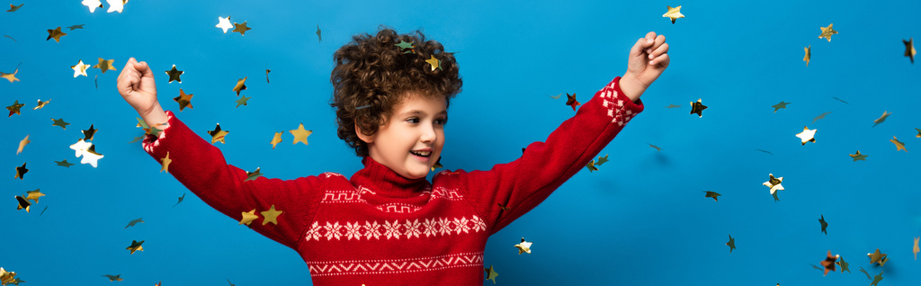 colheita panorâmica de menino animado com as mãos acima da cabeça perto de confete dourado em azul - Foto, Imagem