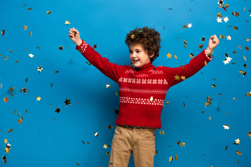 збуджений хлопчик з руками над головою в червоному светрі біля золотої конфетті на синьому
 - Фото, зображення