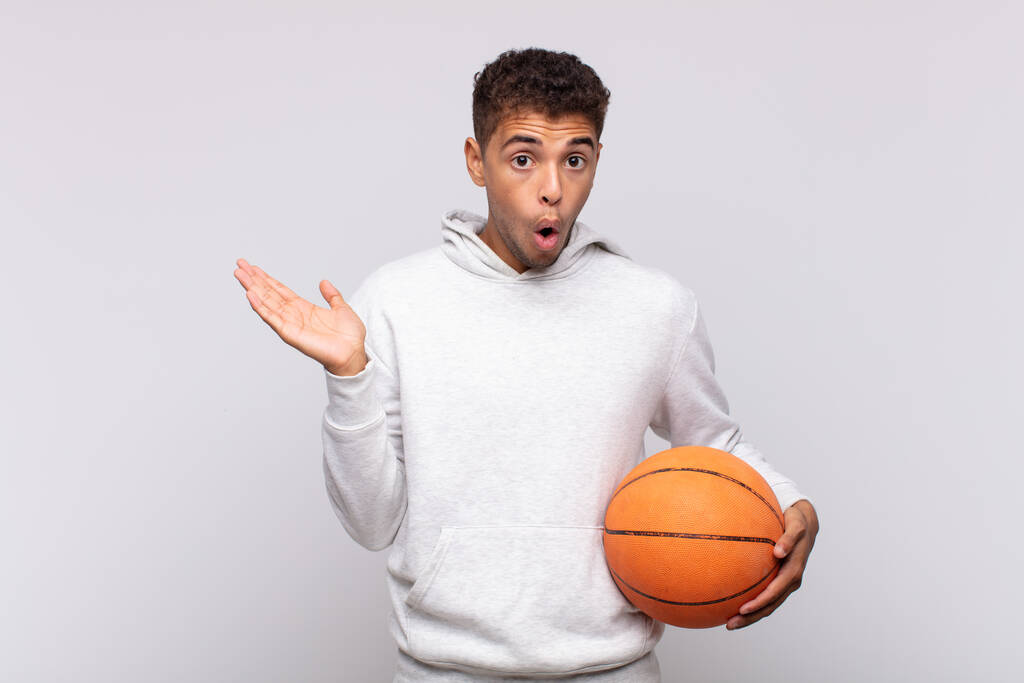 young man looking surprised and shocked, with jaw dropped holding an object with an open hand on the side. basket concept - Photo, Image