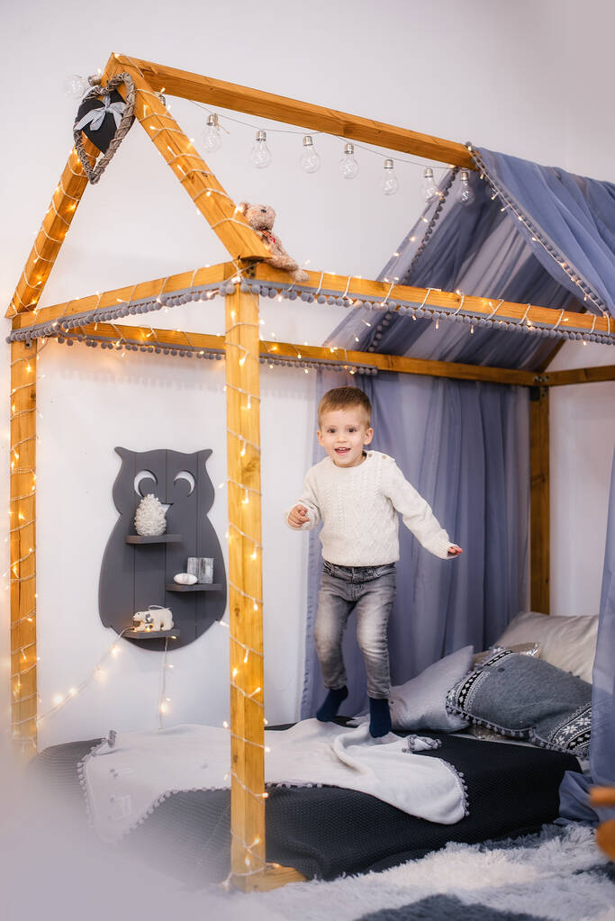 A little boy in a white knitted sweater and jeans jumps merrily and plays on his bed in the form of a tent house, decorated with garlands of lights. Happy childhood. Children room interior. Copy space - Photo, Image