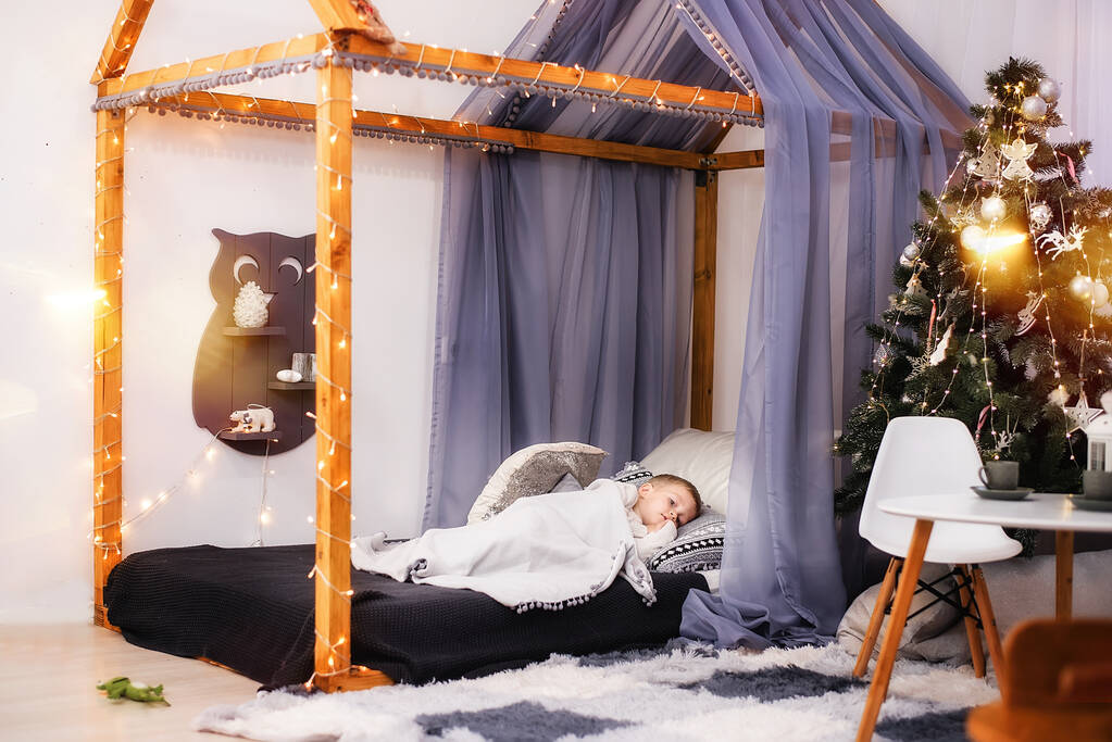 Un petit garçon gît couvert d'une couverture blanche sur son lit en forme de tente, décorée de guirlandes de lumières. Bonne enfance. Chambre d'enfants intérieur. Arbre de Noël. Espace de copie - Photo, image