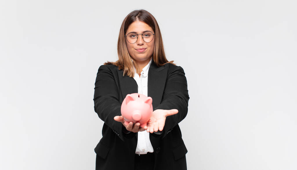 young woman with a piggy bank smiling happily with friendly, confident, positive look, offering and showing an object or concept - Photo, Image