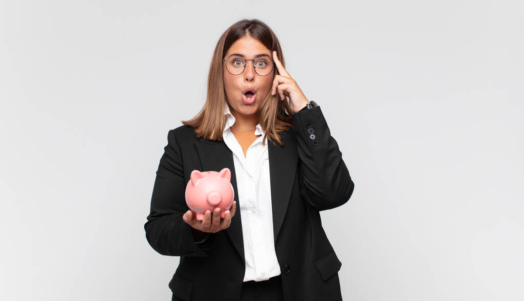 young woman with a piggy bank looking surprised, open-mouthed, shocked, realizing a new thought, idea or concept - Photo, Image