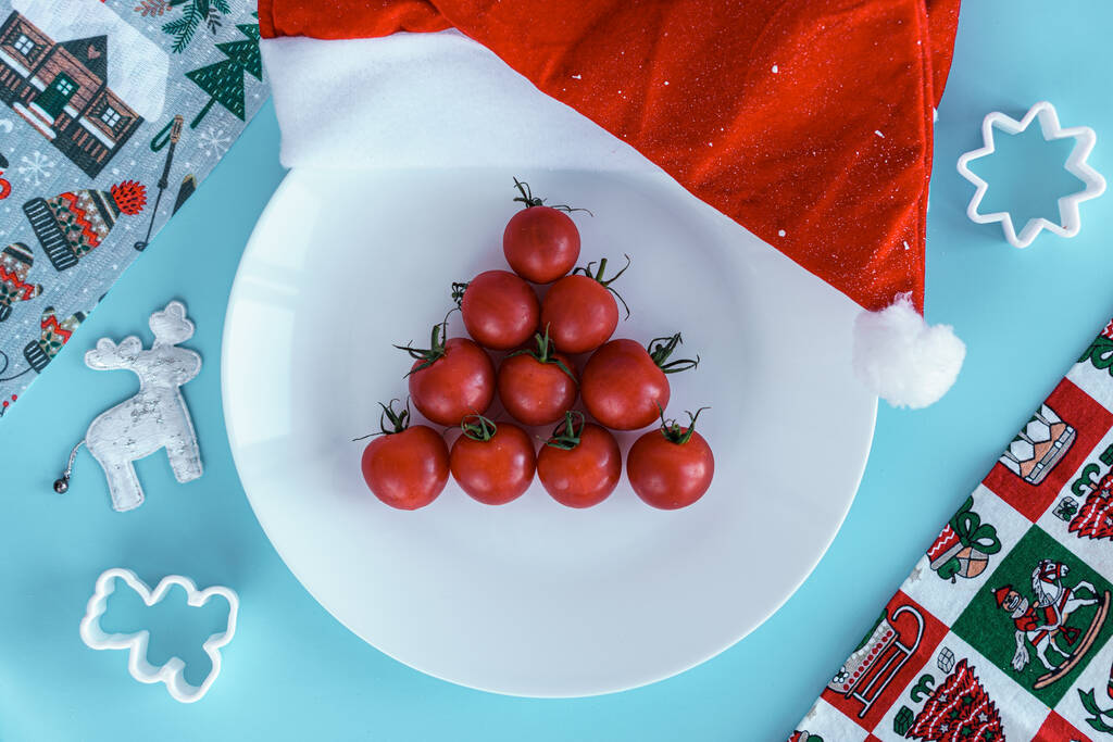 Weihnachten, Winter, Neujahrskonzept - Layout weißer Teller, auf dem rote Kirschtomaten in Dreiecksform mit Weihnachtsmütze und Backwaren auf blauem Hintergrund stehen - Foto, Bild