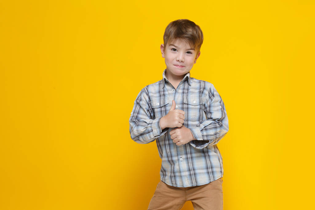 Pequeño niño lindo con una camisa a cuadros muestra el pulgar hacia arriba, aislado sobre un fondo amarillo. - Foto, imagen