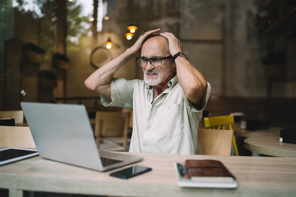 Расстроенный старший мужчина в очках с головой в руках в отчаянии работает удаленно на ноутбуке и смотрит на экран, сидя в кафе - Фото, изображение