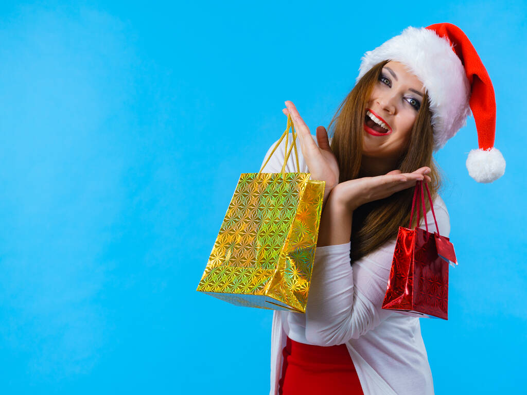 Jonge brunette vrouw geeft graag kerstcadeaus. Vrouw dragen kerstman hoed met cadeautjes feestelijke geschenkzakjes, op blauw - Foto, afbeelding