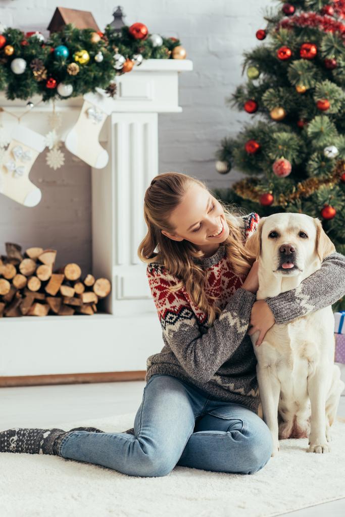 felice donna in maglione seduto sul pavimento e coccole labrador vicino albero di Natale - Foto, immagini