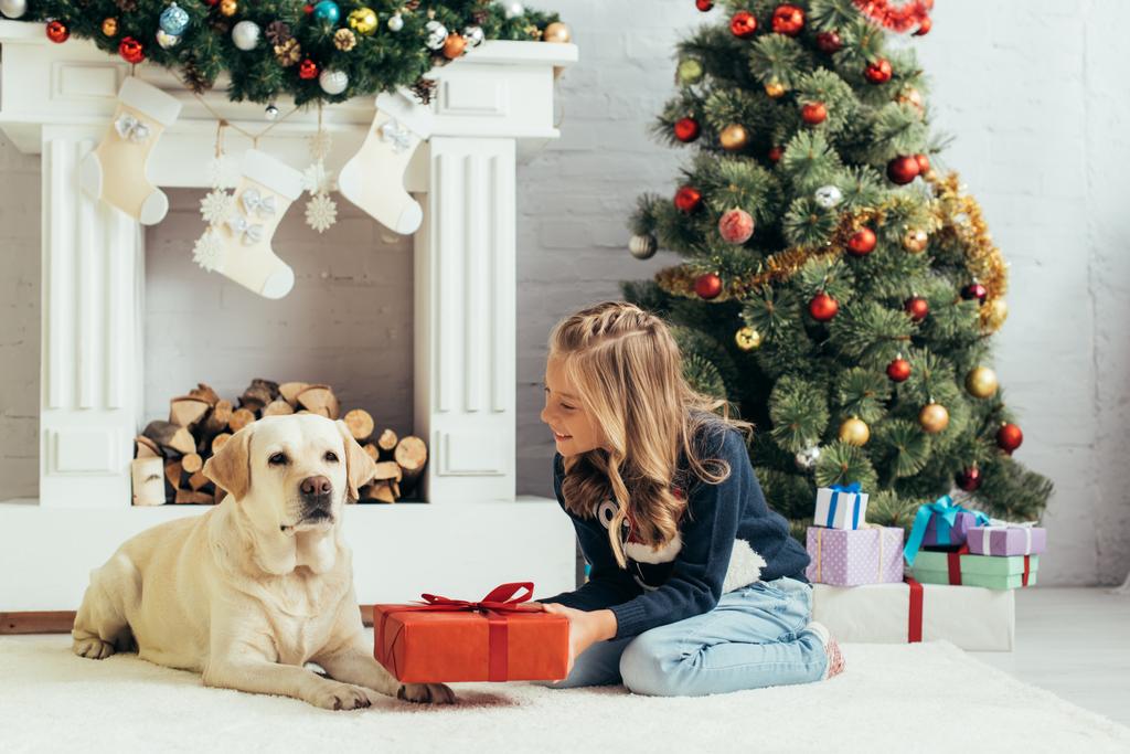 Labrador 'un yanında oturan ve dekore edilmiş oturma odasında hediyesini tutan sıcak süveterli mutlu çocuk, Noel konsepti.  - Fotoğraf, Görsel