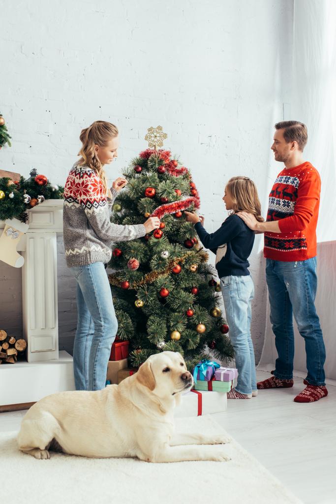Labrador liegt neben glücklicher Familie und schmückt Weihnachtsbaum im Wohnzimmer - Foto, Bild